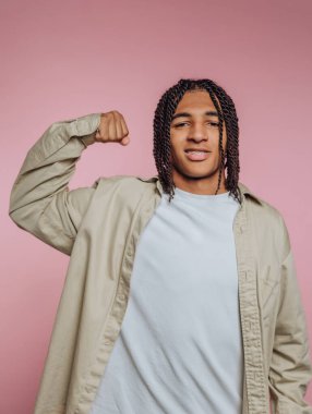 A young man with braided hair smiles and flexes his arm to show off his muscles. He wears a casual shirt and stands against a soft pink backdrop, exuding confidence.