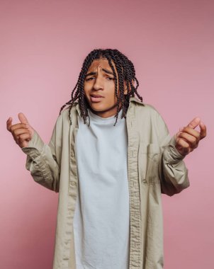 A young individual with braided hair looks puzzled and raises their hands in a questioning manner. This scene occurs in a cozy indoor space with a pink backdrop.