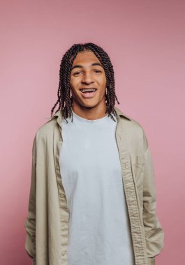 A young individual stands with a joyful expression against a soft pink backdrop. Their braided hair and relaxed outfit convey a sense of casual style and friendliness.