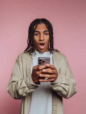 A young man with braided hair shows an expression of surprise while looking at his smartphone. The background is a soft pink, enhancing his astonished look.