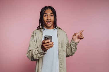 A young individual with braided hair holds a smartphone while expressing excitement. The setting features a vibrant pink backdrop that enhances the lively mood of the scene.