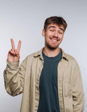 A young man stands against a plain light backdrop, smiling widely while displaying a peace sign with his fingers. He wears a casual shirt over a sweater, radiating positive energy.