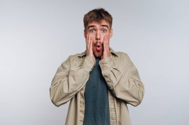 A young man stands with a surprised expression, placing his hands on his cheeks. He has short hair and wears a light shirt over a dark t-shirt in a simple setting.