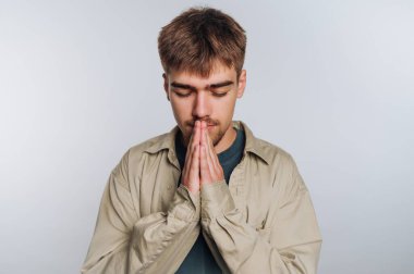 A young man stands in a well-lit indoor space, eyes closed and hands pressed together in a gesture of contemplation or prayer. He appears focused and reflective, wearing a simple outfit.