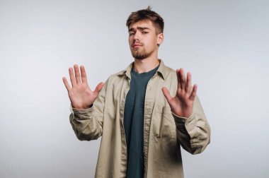 A young man stands with raised hands, showing a gesture of disagreement. He wears a casual shirt and a t-shirt underneath. The background is simple and neutral.