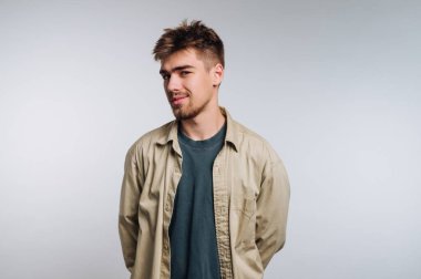 A young man stands confidently with his hands behind his back, wearing a light-colored shirt over a dark t-shirt. The background is neutral and plain, highlighting his expression.