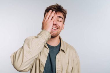 A young man stands in a light studio, smiling with his hand covering part of his face. His outfit is casual, and he appears to be enjoying a light-hearted moment.