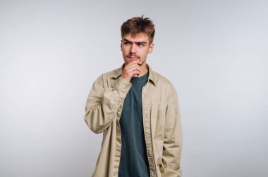 A young man stands in a brightly lit studio, wearing a casual outfit. He has a thoughtful expression as he touches his chin, lost in contemplation about something.
