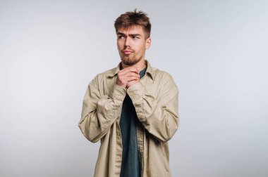 A young man stands in a simple studio, wearing a light beige shirt over a dark shirt. He looks pensive, with his hands clasped together. The background is plain and light.