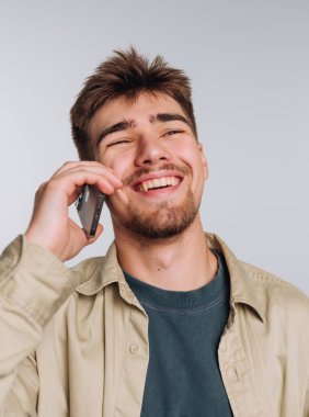A joyful young man enjoys a conversation on his phone, smiling widely in a well-lit space. His casual outfit and relaxed demeanor create a cheerful atmosphere.
