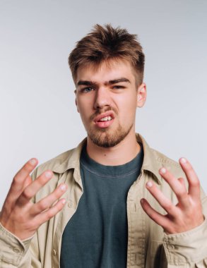 A young man stands in a bright studio, showing a frustrated expression. His hands are raised as if seeking answers, and he wears a casual outfit with a light jacket.