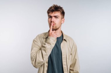 A young man stands in a bright, minimalistic room. He has short, tousled hair and a neutral expression, raising his finger to his lips to indicate silence.