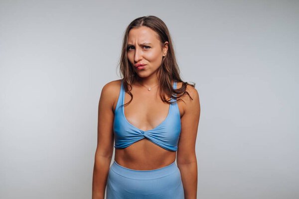 A woman stands confidently in a blue top and matching bottom, playfully expressing her emotions in a minimalist studio. The background is neutral, emphasizing her charm.