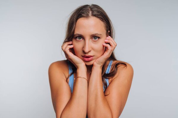 A woman stands with her hands on her face, displaying a look of intensity and vulnerability. The neutral backdrop emphasizes her striking features and emotional expression.