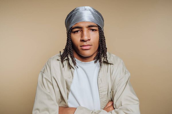 A young man stands with arms crossed, displaying a confident expression. He wears a light shirt and a silver headscarf, set against a beige backdrop that highlights his features and style.