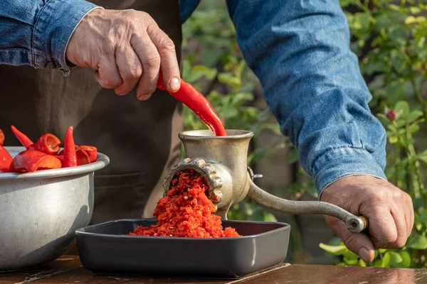 Preparation of homemade sauce with a sweet bell peppers, hot pepper chilli with a grinding machine. Man grinding red peppers on old meat grinder outdoors close-up. Vegetable sauce Healthy organic food concept