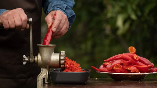 Preparation of homemade sauce with a sweet bell peppers, hot pepper chilli with a grinding machine. Man grinding red peppers on old meat grinder outdoors close-up. Vegetable sauce Healthy organic food concept
