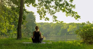 Back view of unrecognizable woman practicing yoga in summer outdoors. Rear view of female sitting in meditation pose on yoga mat in lotus position, raising hands up, doing stretching exercise outside