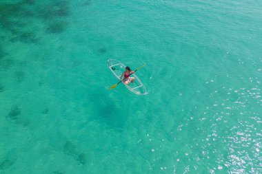 Aerial top view of Asian woman, a tourist, paddling a boat, canoe, kayak or surfboard with clear blue turquoise seawater, Andaman sea in Phuket island in summer season, Thailand. Water in ocean