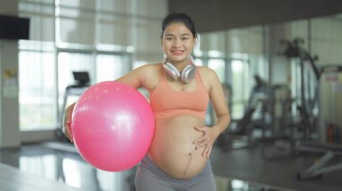 Jimnastik salonunda mutlu Asyalı hamile bir kadının portresi. Bebek sahibi olmak. Antrenman egzersizi. Spor malzemeleriyle içeride egzersiz yap. Atletik. Aile hayatı işte. Anne sevgisi. Rekreasyon.