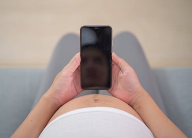 Mutlu Asyalı hamile bir kadının portresi. Evde yatak odasında ekranı olan çevrimiçi cep telefonu teknolojisi kullanıyor. Bebek sahibi olmak. Aile hayatı işte. Annen seni seviyor.