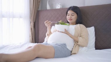 Evdeki yatak odasında diyet yapmak için salata yiyen mutlu Asyalı hamile kadının portresi. Bebek sahibi olmak. Aile hayatı işte. Annen seni seviyor.