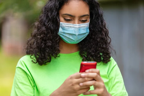 人種混合出生アフリカ系アメリカ人10代の少女顔マスクを着用し コロナウイルスCovid 19パンデミックの間にソーシャルメディアのための携帯電話を使用して若い女性 — ストック写真