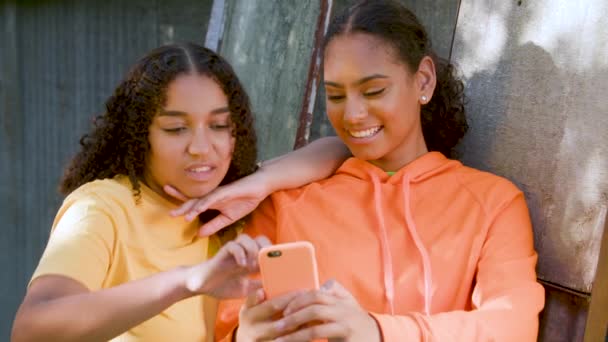 アフリカ系アメリカ人の女の子の10代の若い女性がソーシャルメディアやテキストメッセージのためのスマートフォンや携帯電話を使用して市内の屋外で2つの美しく 笑顔混合レース — ストック動画