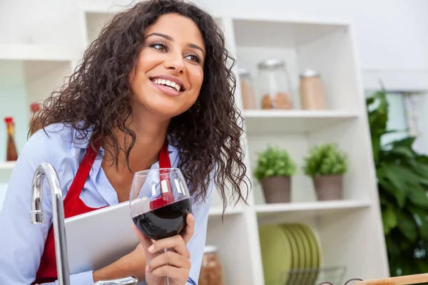 Beautiful young Latina Hispanic woman with perfect teeth smiling, laughing and relaxing using tablet computer while drinking a glass of red wine in her kitchen at home