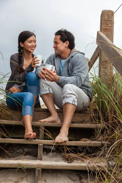 年轻的亚裔男子和女子 男孩和女孩 约会对象坐在木制台阶上俯瞰海滩 喝着茶杯或咖啡 图库图片