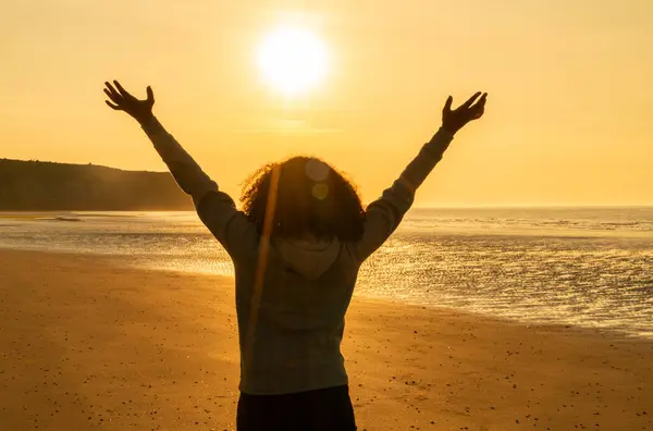 夕阳西下站在荒芜海滩上庆祝的年轻女子举起双臂的后视镜 免版税图库图片
