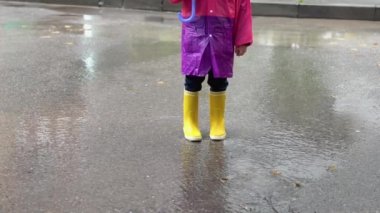 Çocuk yağmurda oyun oynuyor. Şemsiyeli ve çizmeli çocuklar sağanak yağmurda dışarıda oynuyorlar. Çamurlu su birikintisine atlayan küçük çocuk. Çocuklar yağmurlu sonbahar havasına göre eğlencelidir. Tropikal fırtınada koşan çocuk.