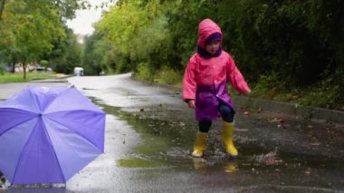 Şemsiyeli ve çizmeli çocuklar sağanak yağmurda dışarıda oynuyorlar. Çocuk yağmurda oyun oynuyor. Çamurlu su birikintisine atlayan küçük çocuk. Fırtınada koşan çocuk