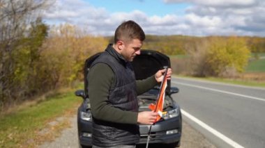 Genç adam diğer yol kullanıcılarını uyarmak için kırmızı üçgen hazırlıyor, araba arızası veya motor arızası kırsal caddede duruyor.