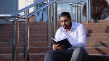 Mutlu Hintli öğrenci merdivenlerde oturur bir üniversite kampüsünde kitap okur.