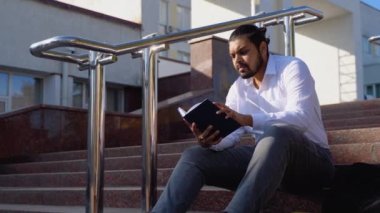 Hintli öğrenci merdivenlerde oturur bir üniversite kampüsünde kitap okur..