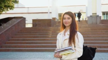 Neşeli öğrenci kız okul binasının yakınlarında sırt çantasıyla poz veren ve kameraya gülümseyen kitaplara sarılıyor. Modern eğitim, üniversite harcı ve bağışlar