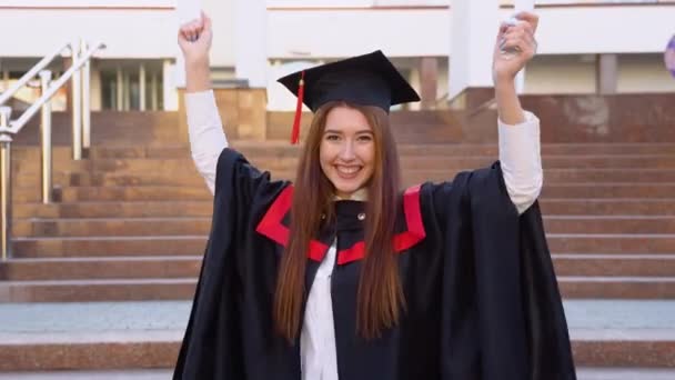 快乐的女大学生站在一个硕士的衣橱里 得意洋洋地在他头上挥动着一张文凭 — 图库视频影像