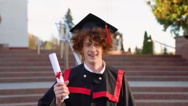 穿着毕业典礼服装的快乐的家伙在镜头前展示他的文凭和微笑 — 图库视频影像