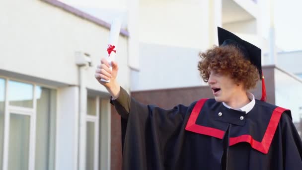 一个得意洋洋的男性毕业生高举文凭站在大学近旁的低角度画像 从下面这位年轻英俊的男人为自己在庆祝大学毕业典礼上取得的学术成就而自豪 — 图库视频影像