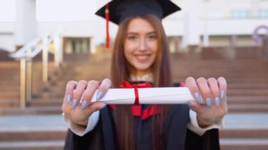 Üniversite mezunu Master Mast 'ta duruyor ve kameranın önünde diplomasını tutuyor. Kameranın odak noktası parşömen.