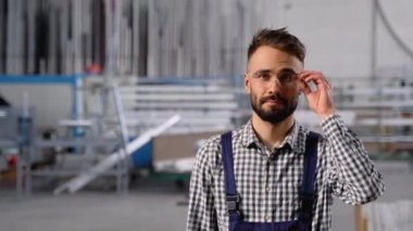 Sakallı, koruyucu gözlüklü, atölyede dikilirken kameraya bakan fabrika işçisinin portresi..