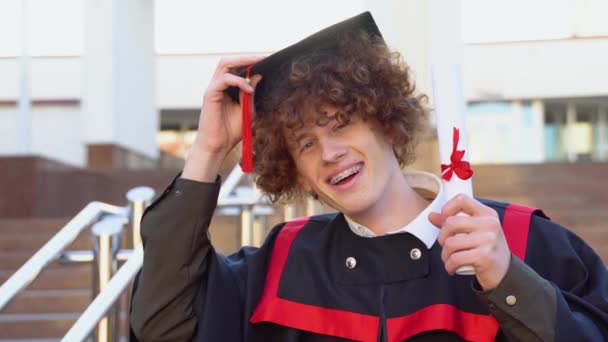 ブレースを持つ赤髪巻き男はマスターマントルに立っており 帽子と卒業証書を保持しています — ストック動画