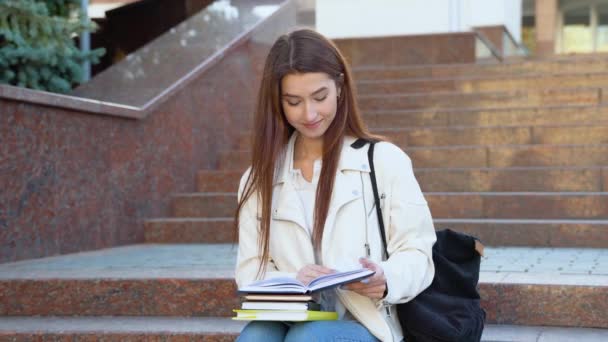 若い女性学生は大学の建物の近くで本を読んでいる 女の子は勉強していて 大学のキャンパスで試験の準備をしています 教育の概念 高校生活 — ストック動画