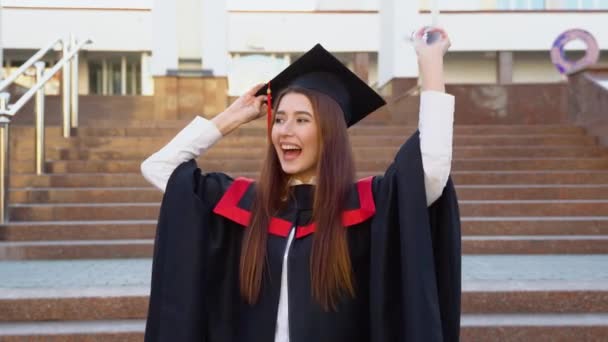 一位女大学生站在硕士的衣橱里 戴着硕士的帽子 得意洋洋地在头上挥动着毕业证书 — 图库视频影像