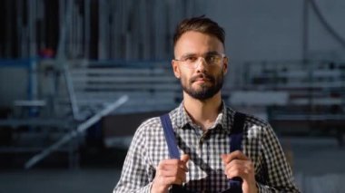 Sakallı fabrika işçisinin belini kaldırıp atölyede dikilirken kameraya bakıyor.