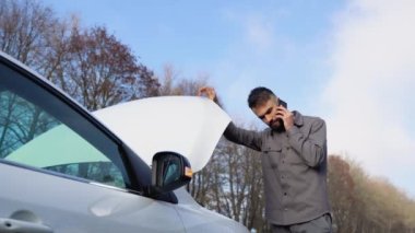 Üzgün şoför bozuk arabanın yanında duruyor, cep telefonuyla acil servisi arıyor ve akıllı telefondan konuşuyor. Çaresiz genç bir adam kaza geçirdi..