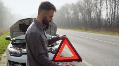 Genç adam diğer yol kullanıcılarını uyarmak için kırmızı üçgen hazırlıyor, araba arızası veya motor arızası kırsal caddede duruyor