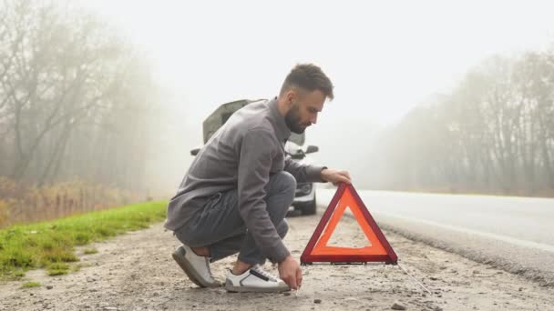 男は壊れた車の近くの霧の道に三角形の注意標識を置く 旅行の概念 — ストック動画