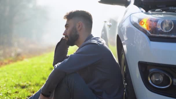 若い男は自動車が故障したので携帯電話で話している車の近くに座っている 白人男性は友人に車の修理を手伝おうとしています — ストック動画